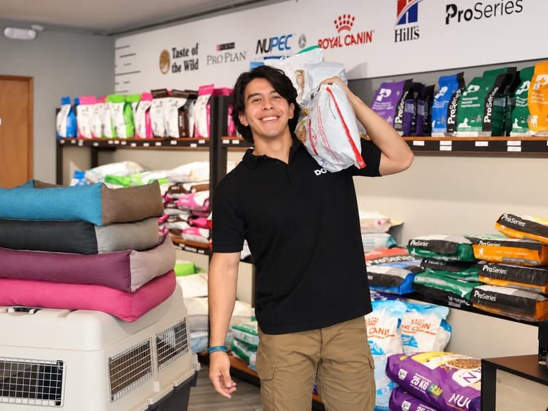 Colaborador de DOGO sonriendo con un costal de croquetas al hombro, junto a camas para perro, una transportadora y anaqueles de ProSeries y Royal Canin